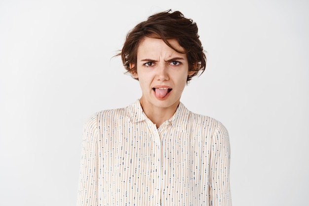 Free photo portrait of childish young woman express aversion and dislike by showing tongue and frowning, looking at something disgusting, bad taste, standing on white wall