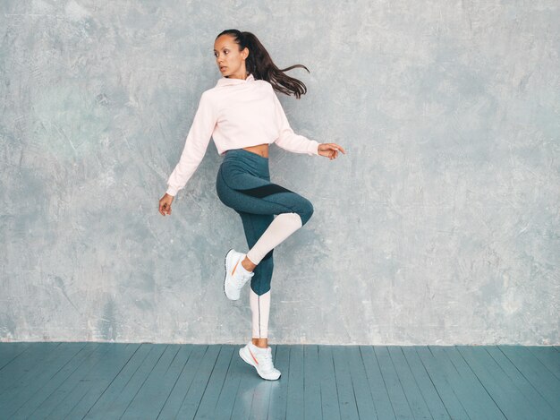 Portrait of confident fitness woman in sports clothing looking confident.Female jumping in studio near gray wall