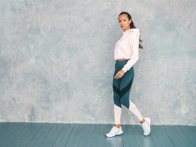 Portrait of confident fitness woman in sports clothing looking confident.Female walking in studio near gray wall