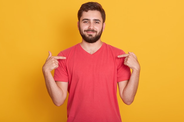 Free photo portrait of confident good looking caucasian man wearing red casual t shirt, standing over yellow, pointing on his t shirt