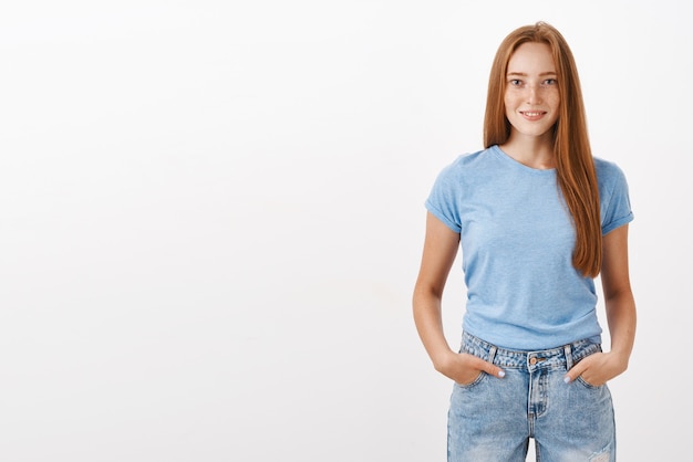 Portrait of confident good-looking ginger girl with cute freckles in blue t-shirt and jeans holding hands in pockets and smiling with self-assured expression