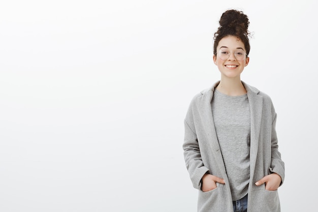 Portrait of confident happy good-looking girl with curly hair combed in bun, wearing gray coat and glasses, holding hands in pockets and smiling cheerfully