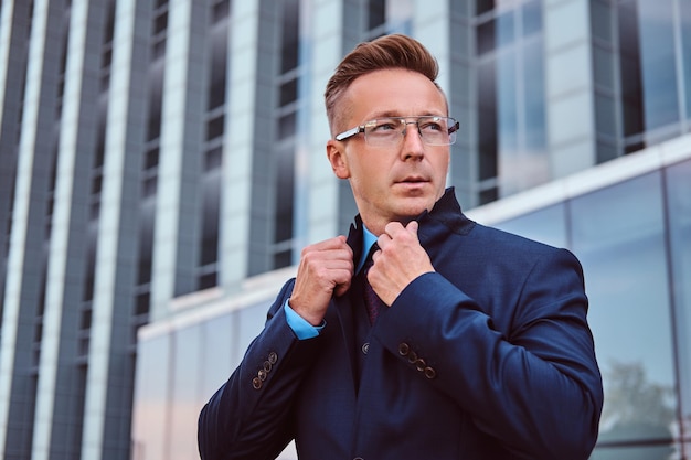 Portrait of a confident man in elegant suit and glasses correct his jacket while standing against a skyscraper background.