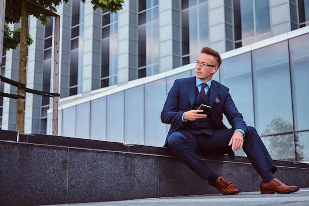 Free photo portrait of a confident stylish businessman dressed in an elegant suit holds a smartphone and looking away while sitting outdoors against skyscraper background.