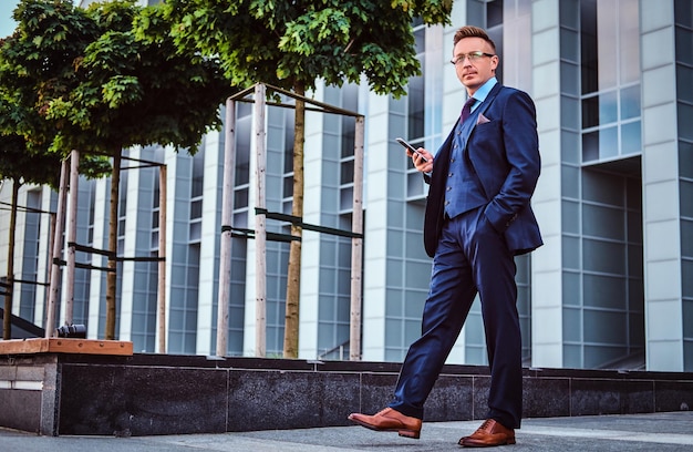 Free photo portrait of a confident stylish businessman dressed in an elegant suit holds a smartphone and looking away while standing outdoors against skyscraper background.