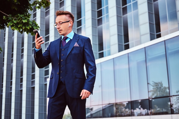 Free photo portrait of a confident stylish businessman dressed in an elegant suit holds a smartphone and looking away while standing outdoors against skyscraper background.