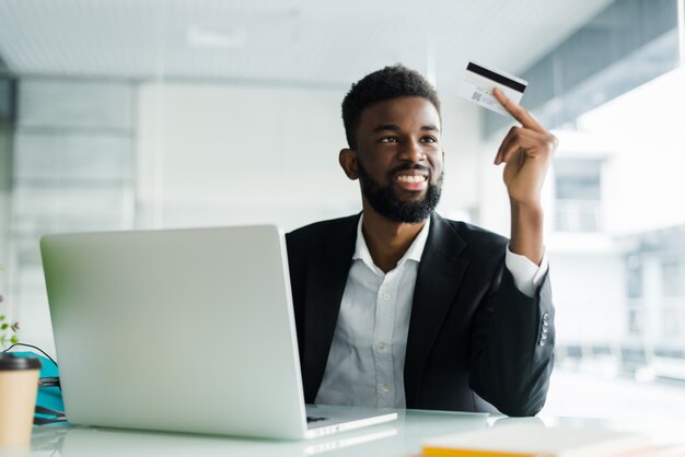 Portrait of confident young african man holding credit card with laptop paying via internet