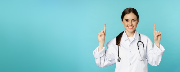 Free photo portrait of confident young woman doctor medical worker in coat pointing fingers up and smiling showing medicine advertisement clinic promo toquoise background