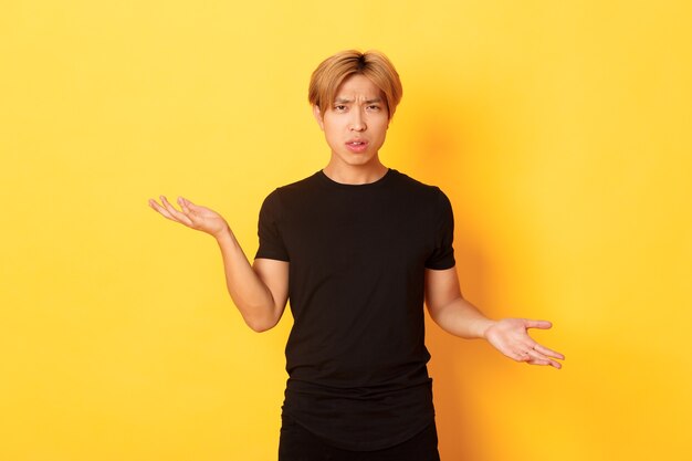 Portrait of confused and annoyed asian guy spread hands sideways, cant understand something, standing yellow wall