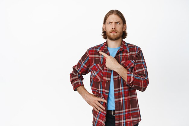 Portrait of confused blond man with beard, frowning suspicious, pointing finger and looking left displeased, clueless about smth, standing against white background.