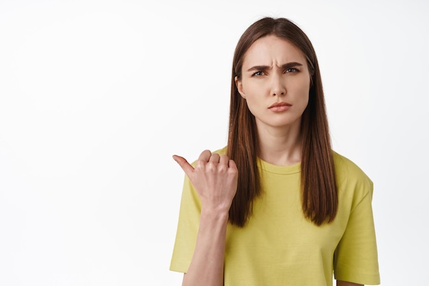 Portrait of confused brunette woman pointing left, squinting and frowning puzzled, showing smth strange, asking question, what is that gesture, white background