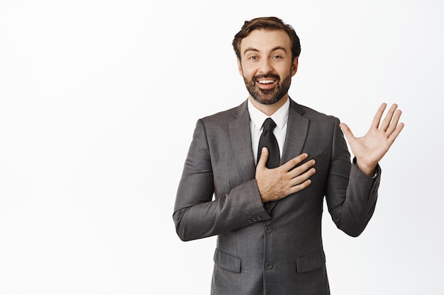 Free photo portrait of corporate man raising palm put hand on heart and smiling sincere making promise introduce himself standing in grey suit over white background