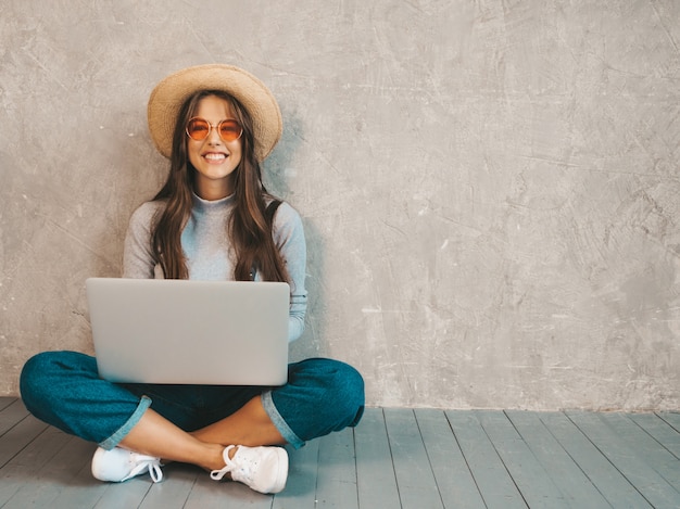 Portrait of creative young smiling woman in sunglasses. Beautiful girl sitting on the floor near gray wall. 