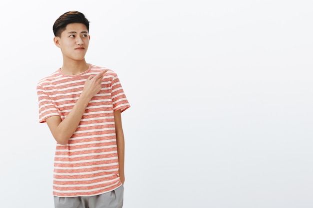 Free Photo portrait of curious nice young asian male model in striped t-shirt standing relaxed over grey wall with hand in pocket looking and pointing at upper right angle seeing interesting copy space