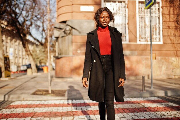 Portrait of a curly haired african woman wearing fashionable black coat and red turtleneck walking on zebra crossing