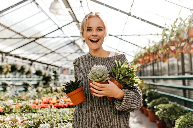 Portrait of curly woman in gray sweater holding lot of potted plants. Green-eyed blonde with smile poses at plant store.