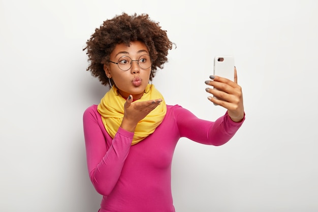 Portrait of cute African American woman sends air kiss, takes selfie via smartphone, wears transparent glasses, makes rounded lips, wears vivid clothes, isolated over white wall.