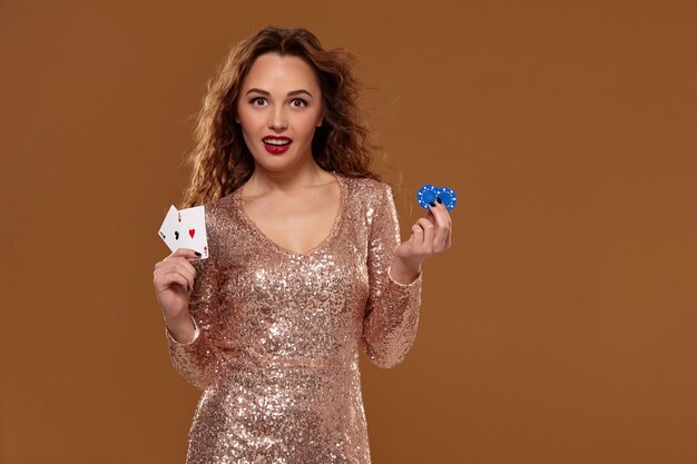 Portrait of cute brown-haired caucasian girl on brown background in golden cocktail dress at casino holds pair of aces and chips in her hands. Gambling industry, poker, casino