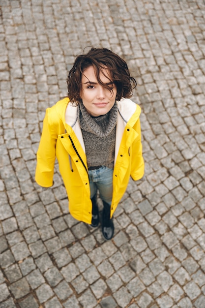 Free Photo portrait of cute female in yellow stylish coat looking up on camera while standing on paving stones during her walk