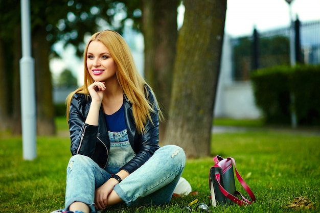 portrait of cute funny blond modern sexy urban young stylish smiling woman girl model in bright modern cloth outdoors sitting in the park in jeans with pink bag