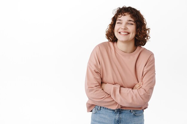 Portrait of cute girl laughs and smiles, standing relaxed in casual pose, cross arms on chest as if feeling chilly, looking aside at upper left corner, standing on white.