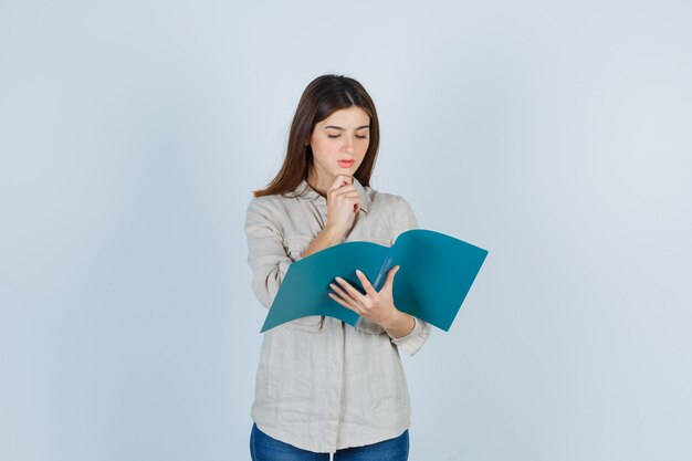 Portrait of cute girl looking through folder, propping chin on finger in shirt and looking pensive