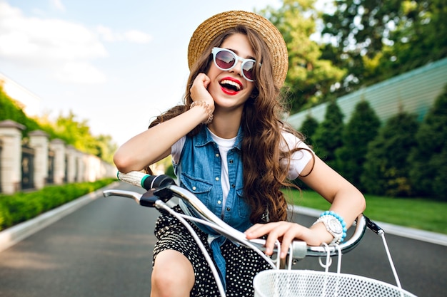 Free photo portrait of cute girl with long curly hair in hat  driving a bike on road. she wears long skirt, jerkin, blue sunglasses. she is smiling to camera.