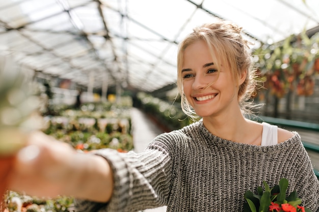 Free photo portrait of cute green-eyed woman with smile poses in greenhouse and holds plant.