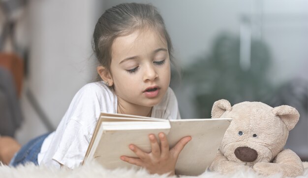 Portrait of a cute little girl reading a book at home, lying on the floor with her favorite toy.