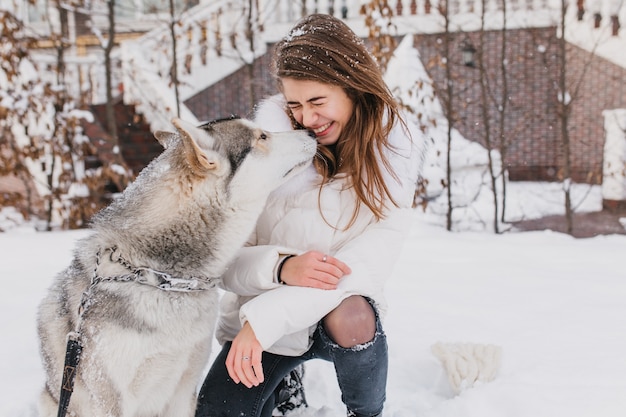 Free Photo portrait cute lovely moments of husky dog kissing fashionable young woman outdoor in snow. cheerful mood, winter holidays, snow time, real friendship, animals love.