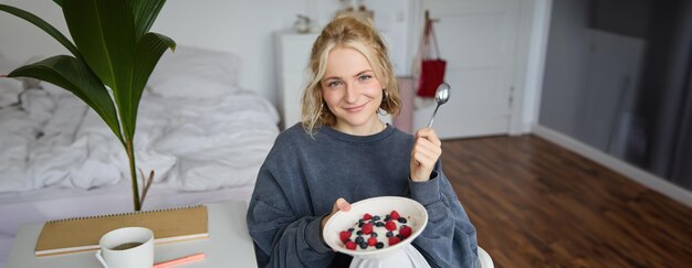 Portrait of cute smiling blond woman eats breakfast in her bedroom looking at camera holding bowl