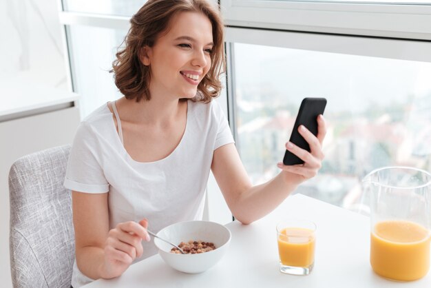 Portrait of cute smiling woman in white tshirt chatting on smartphone while sitting and eating cornflakes at the kitchen table