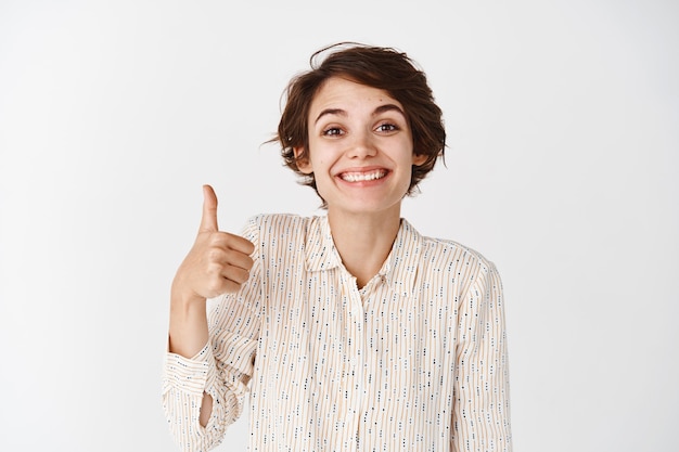 Free Photo portrait of cute supportive girl showing thumbs up and smiling proud, praising you, showing well done, excellent gesture, standing satisfied on white wall