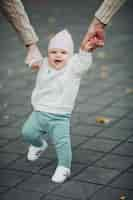 Free photo portrait of cute sweet baby during walking outside parents holding their satisfied child by hands while it walking happy smiling infant in white sweater green pants and white sneakers doing steps