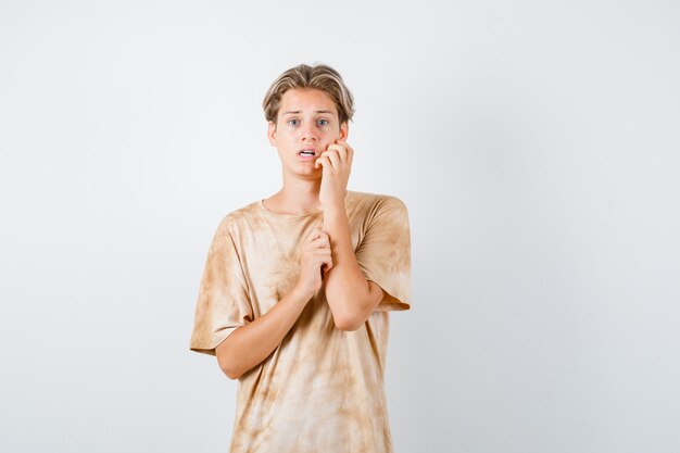 Portrait of cute teen boy keeping hand on cheek in t-shirt and looking frightened front view