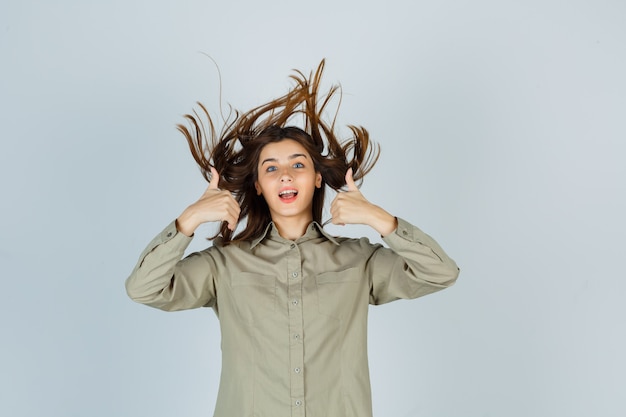 Free photo portrait of cute young female showing double thumbs up while posing with flying hair in shirt and looking blissful front view