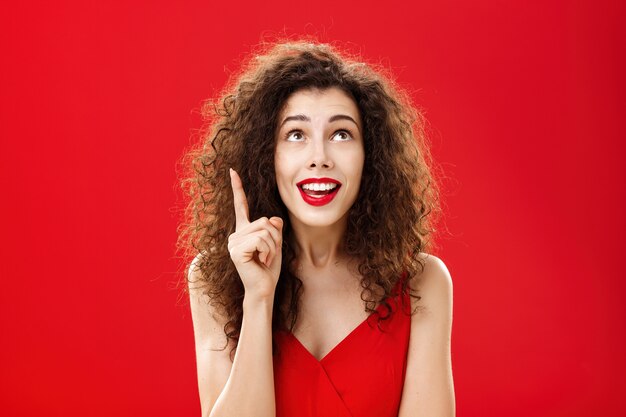 Portrait of delighted charming silly female with curly hairstyle in dress raising index finger in eureka gesture smiling happily looking up having great plan or idea posing charismatic over red wall.