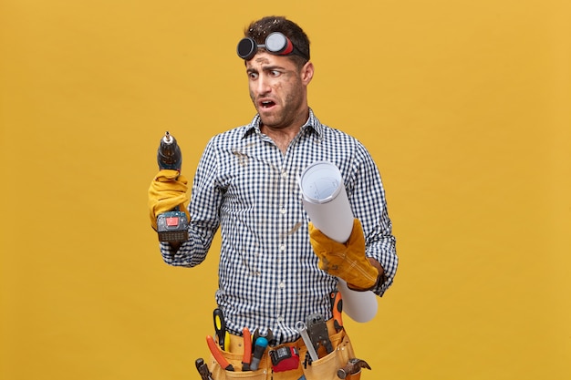 Portrait of dirty repairman having tool belt holding blueprint and drill machine looking at it with anger as it works badly. Craftsman expressing his dissatisfaction with tools and instruments