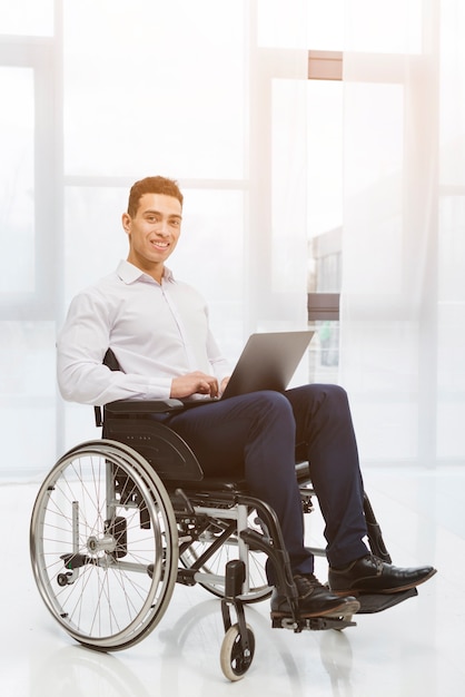 Portrait of a disabled smiling young businessman sitting on wheelchair using laptop in the office