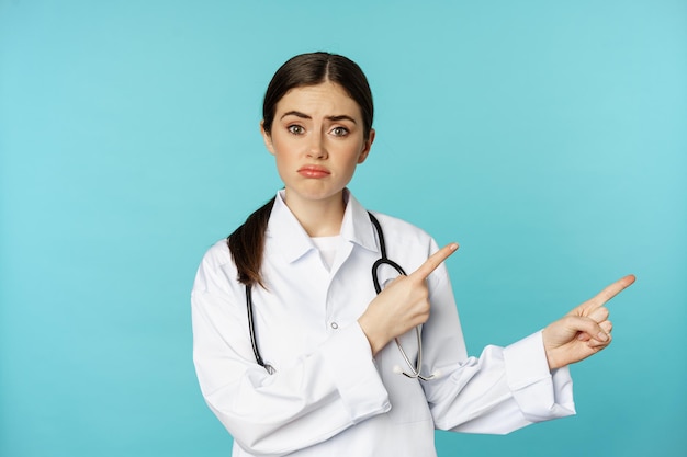 Free photo portrait of disappointed doctor woman medical worker pointing fingers right and looking sad regret g...