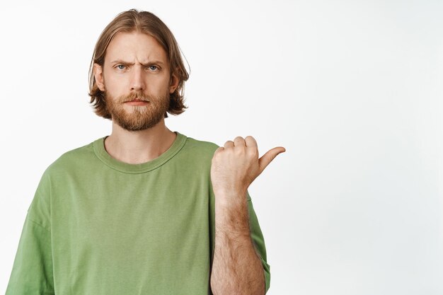 Portrait of disappointed grumpy blond guy pointing right, complaining, staring displeased at camera and frowning, showing smth bad, standing over white background