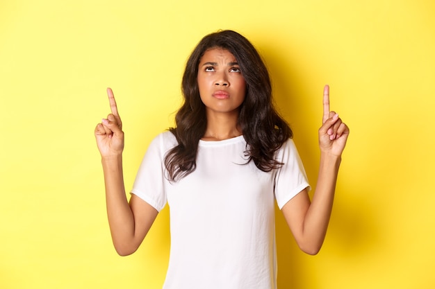 Free photo portrait of disappointed and sad africanamerican girl pointing and looking up frowning displeased