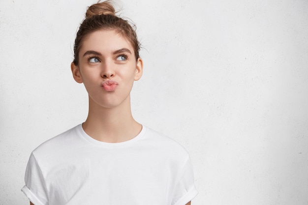 Free photo portrait of discontent pensive female with blue eyes pouts lips and looks thoughtfully upwards, dressed in casual t shirt, isolated over white background. people, emotions, facial expressions concept