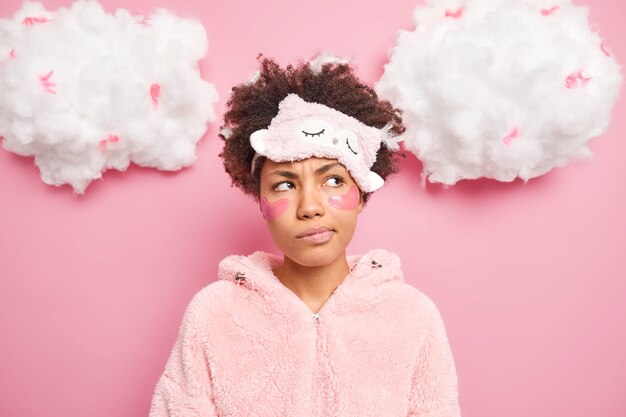 Portrait of discontent thoughtful curly woman looks pensively aside smirks face applies patches under eyes wears sleepmask nightwear isolated over pink wall