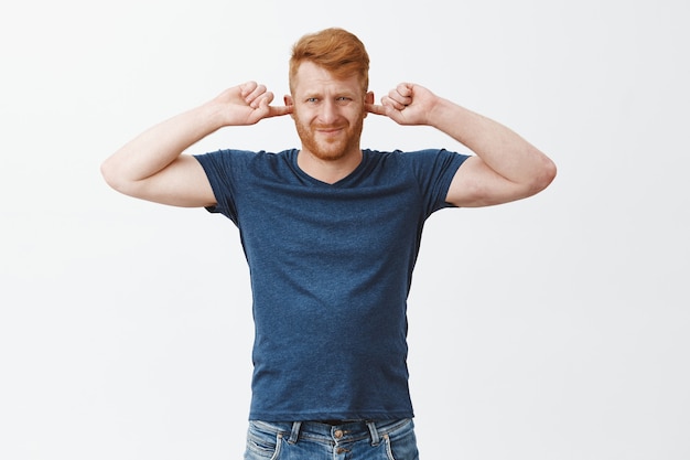 Portrait of displeased attractive strong european man with ginger hair, covering ears, frowning and pursing lips from discomfort, being annoyed with loud sound