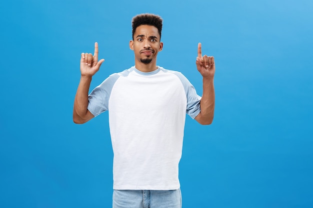 Free photo portrait of displeased intense and doubtful african american male model in casual t-shirt smirking and frowning from disappointment and doubt pointing up seeing something suspicious over blue wall