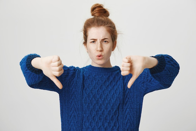 Portrait of displeased unimpressed attractive redhead girl in casual winter sweater showing thumbs down