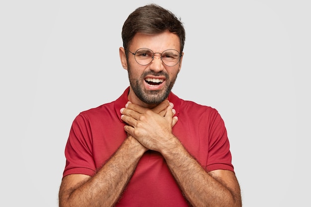 Portrait of displeased young male, feels terrible pain, suffers from sore throat, keeps hands on neck, looks with discontent expression, has problems with health, isolated over white wall