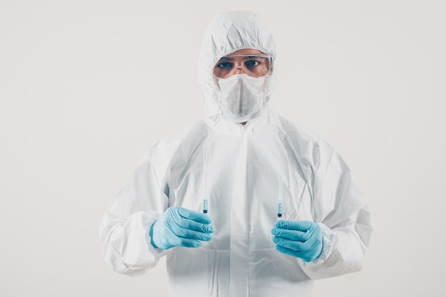 Portrait of a doctor at light background holding 2 syringes with medicine in medical gloves and protective suit