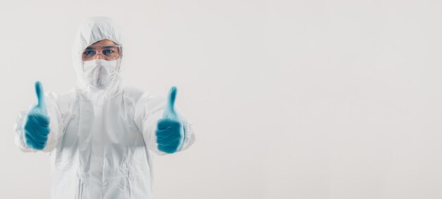 Portrait of a doctor at light background standing and showing thumbs up in mask, medical gloves and protective suit space for text